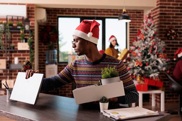 Desempleado deprimido siendo despedido en la víspera de navidad, reuniendo pertenencias para dejar el trabajo en la oficina con decoraciones festivas de navidad. Sentirse triste y molesto por el fracaso profesional y la pérdida del trabajo.