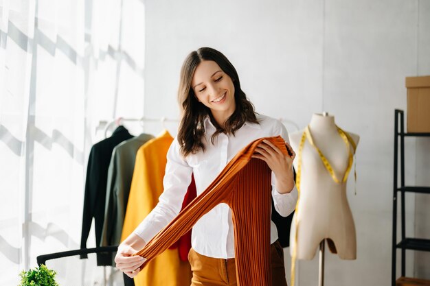 Desenhista de moda mulher falando smartphone e usando laptop com computador tablet digital em estúdio moderno as roupas penduradas nas prateleiras xA