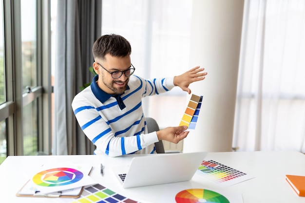 Desenhista homem alegre tendo videoconferência com clientes sentados na mesa na frente do computador segurando paletas de cores gesticulando e sorrindo espaço de cópia