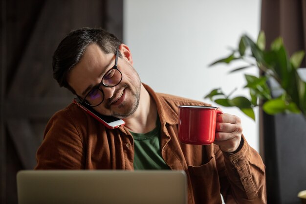 Desenvolvedor cansado trabalhando com laptop, tomando café e ligando do celular em casa