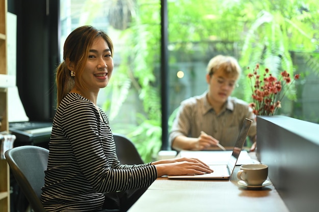 Desenvolvedor de sites feminino confiante sentado no escritório moderno com seu colega e sorrindo para a câmera