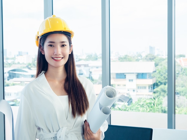 Desenvolvedora de engenheira asiática feliz usando capacete de segurança amarelo segurando papéis de projeto no vidro da janela