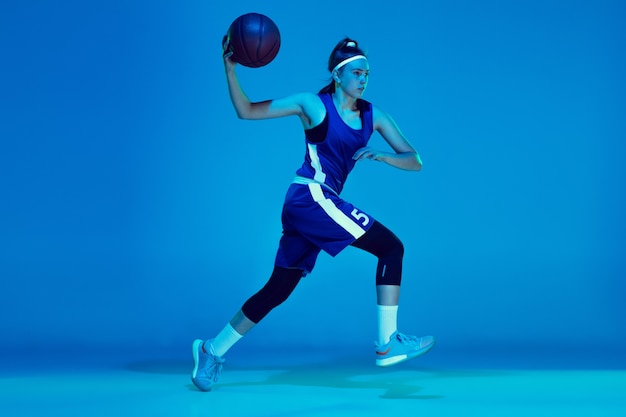 Deseo de ganar. Formación de jugador de baloncesto femenino caucásico joven, prcticing con balón aislado sobre fondo azul en luz de neón. Concepto de deporte, movimiento, energía y estilo de vida dinámico y saludable.