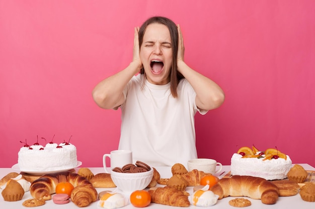 Desespero chocado, mulher irritada vestindo camiseta branca sentada à mesa festiva com vários doces caseiros mantém a dieta cobrindo os ouvidos com as mãos gritando isoladas sobre fundo rosa