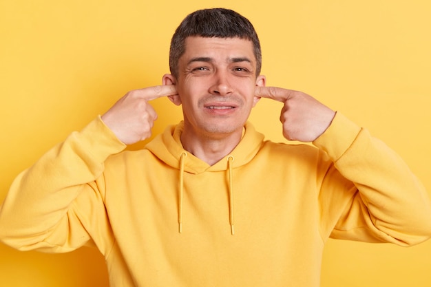 Desespero homem triste vestindo capuz casual cobrindo a boca com os dedos ouvindo barulho alto rosto carrancudo posando isolado sobre fundo amarelo