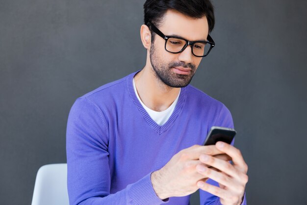 Desfrutando de boas mensagens. jovem bonito segurando um telefone celular enquanto está sentado contra um fundo cinza