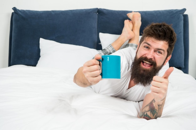 Desfrutando de café fresco homem barbudo feliz hipster beber café da manhã homem sonolento brutal no quarto macho maduro com barba de pijama na cama dormindo e acordado energia e cansaço café fresco