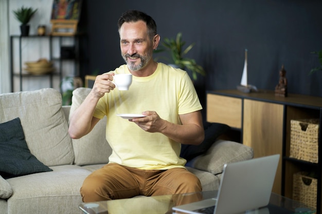 Desfrutando de café ou chá, homem de negócios de meia idade faz uma pausa enquanto trabalha no laptop, sentado no sofá no interior de casa, distância de trabalho, homem de TI maduro bebe café enquanto trabalha no computador