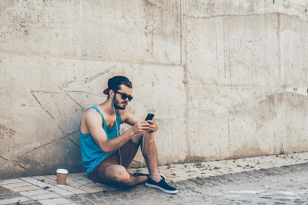 Desfrutando de música ao ar livre. Jovem bonito usando fones de ouvido, segurando um telefone inteligente e olhando para ele enquanto está sentado ao ar livre e encostado na parede de concreto