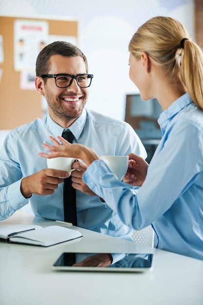 Desfrutando de ótimas relações de trabalho. Dois jovens felizes em trajes formais segurando xícaras de café e discutindo algo enquanto trabalham juntos
