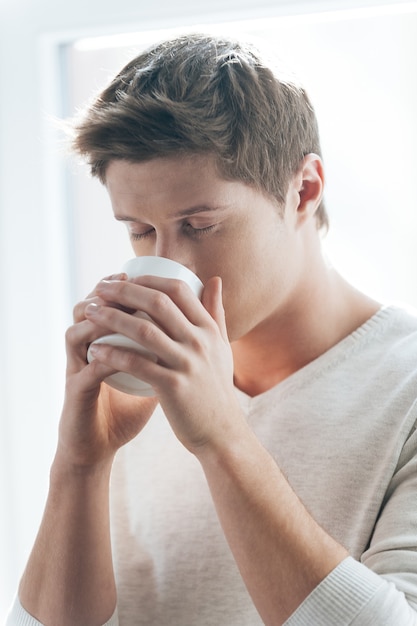 Desfrutando de um café fresco. Jovem bonito tomando café em pé contra a janela