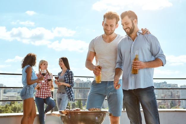 Desfrutando de um churrasco juntos, dois jovens sorridentes, bebendo cerveja e assando carne na grelha