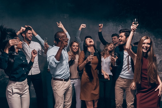 Desfrutando de uma festa incrível. Grupo de jovens lindos dançando com taças de champanhe e parecendo felizes