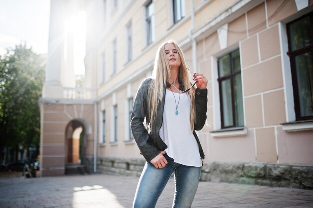 Desgaste de mulher loira elegante em jeans, óculos escuros e jaqueta colocada na rua na luz do sol