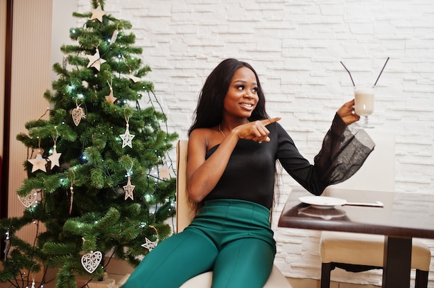 Desgaste de la modelo afroamericana delgada de la belleza en la blusa negra y los pantalones largos verdes de las piernas posó en el café y bebe el café con leche contra el árbol de año nuevo. Humor navideño.