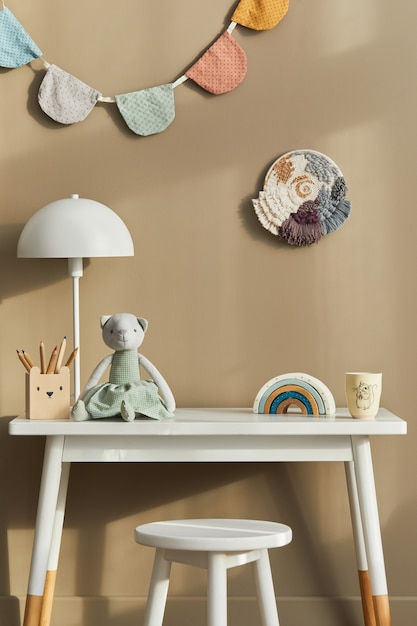 Design de interiores de um elegante quarto infantil com mesa branca, brinquedos de madeira, acessórios infantis, lâmpada branca, decoração aconchegante e bandeiras de algodão penduradas na parede bege.