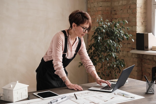 Designer de arquitetura feminina concentrada em óculos em pé na mesa com planta e edição de arquivo no laptop