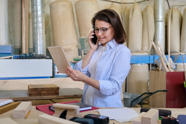 Designer de carpinteiro feminino trabalhando na oficina de carpintaria, escolhendo materiais, falando ao telefone. Negócio industrial, marcenaria