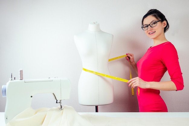 Designer de costureira linda garota com óculos mede a máquina de costura manequim e fundo branco