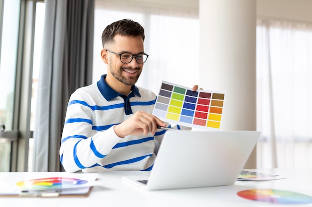 Designer de homem alegre tendo videoconferência com clientes sentados na mesa em frente ao computador segurando paletas de cores gesticulando e sorrindo espaço de cópia