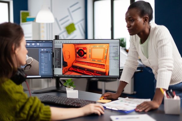 Designer de jogos femininos multiétnicos olhando para um computador com dois monitores trabalhando juntos em um projeto no escritório do estúdio