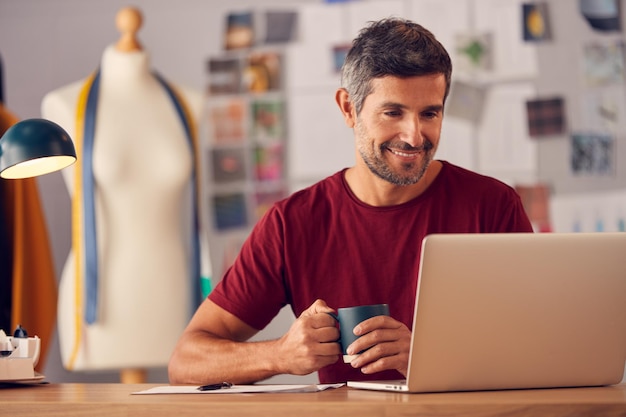 Designer de moda masculino maduro em estúdio sentado na mesa trabalhando no laptop