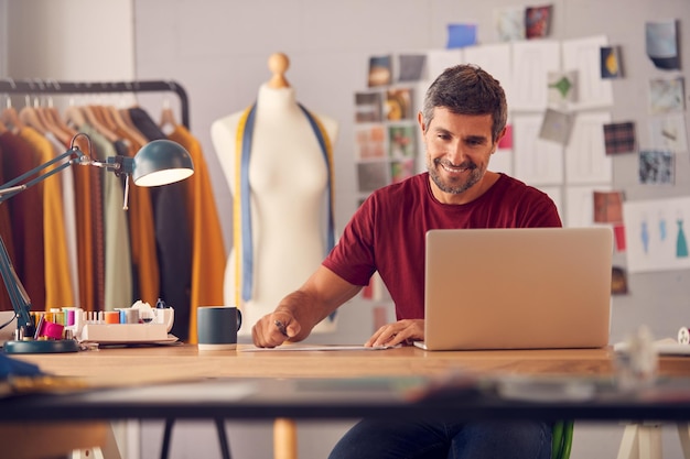 Designer de moda masculino maduro em estúdio trabalhando em esboços ou documentos na mesa com laptop