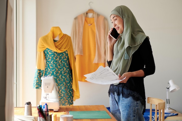 Designer de moda muçulmano alegre segurando esboços e falando ao telefone com o cliente