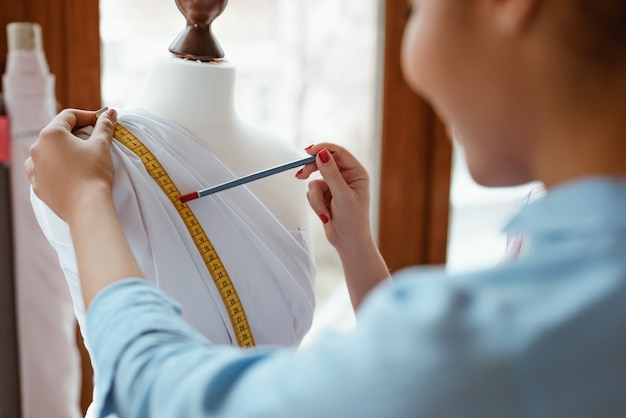 Designer de moda no trabalho. jovem mulher africana, alfaiate está olhando para o manequim com vestido branco. ela medindo roupas