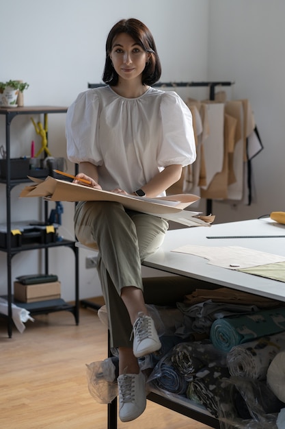 Designer de roupas de esgoto jovem vestida com roupas da moda, sentar na mesa no ateliê de ateliê