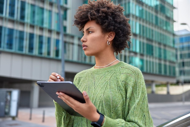 Designer feminina talentosa com stylus e tablet desenha esboços ou adesivos para redes sociais enquanto caminha ao ar livre em local urbano usa jumper verde casual smartwatch para trabalho à distância