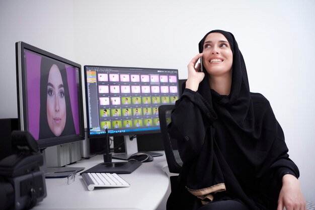 Designer gráfico feminino muçulmano falando ao telefone. Freelancer editando fotos em dois monitores. Garota hijab no local de trabalho