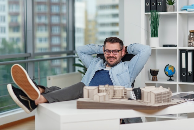 Designer masculino barbudo confiante de óculos sentado em uma pose relaxada no estúdio de design satisfeito com seu projeto