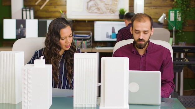 Designer trabalhando em um laptop e arquiteto, observando projetos no interior de um escritório moderno