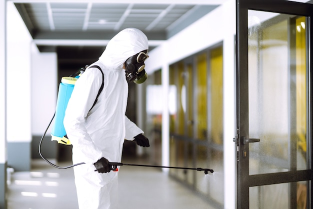 Desinfecção de escritório para prevenir COVID-19, Homem em traje de proteção de materiais perigosos com produtos químicos em spray.