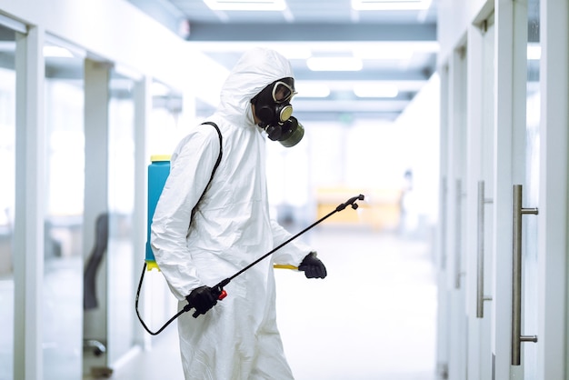 Desinfecção de escritório para prevenir COVID-19, Homem em traje de proteção de materiais perigosos com produtos químicos em spray.