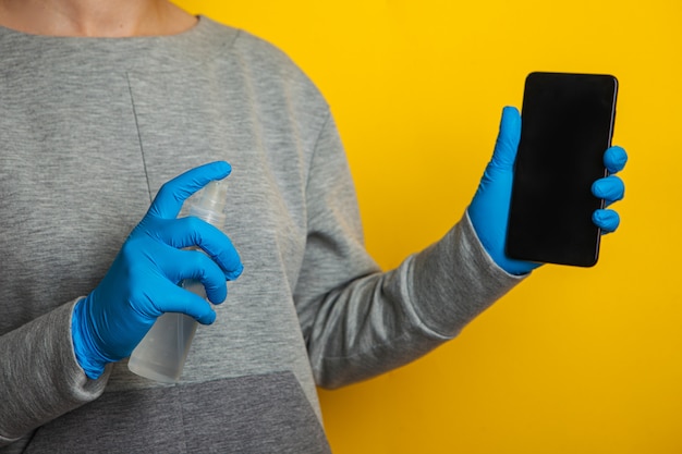 Desinfecção do telefone. Uma mulher com uma máscara médica e luvas de borracha tem um telefone e um desinfetante em spray nas mãos.