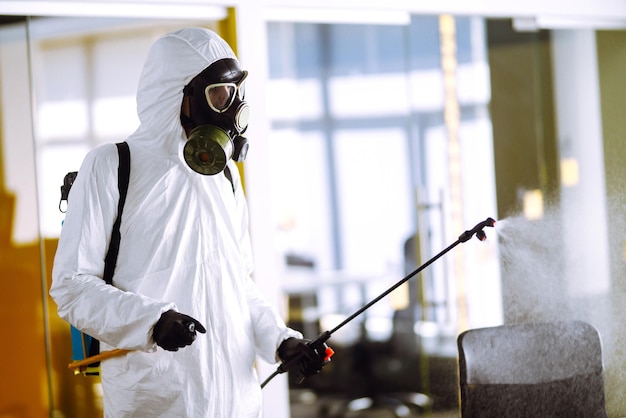 Desinfección de la oficina para prevenir COVID19 Hombre con traje protector de materiales peligrosos con productos químicos en aerosol