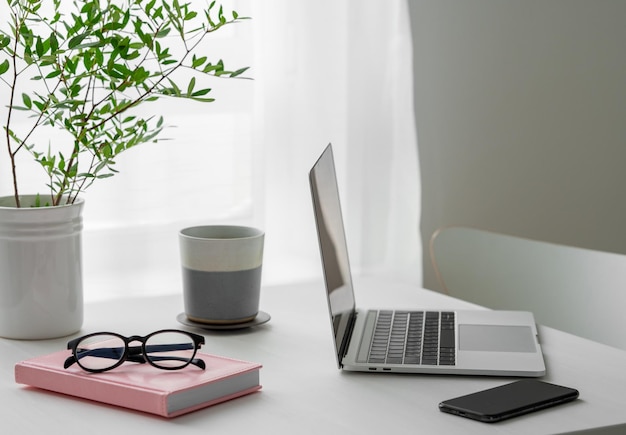 Desktop com laptop, xícara de café, bloco de notas de telefone e óculos na frente de uma janela com a luz do sol da manhã
