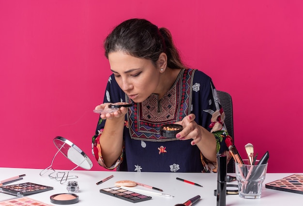 Desorientada joven morena sentada a la mesa con herramientas de maquillaje sosteniendo y oliendo rubor aislado en la pared rosa con espacio de copia