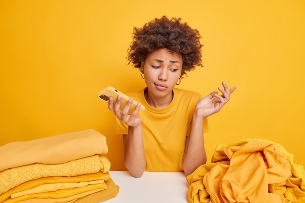 Desorientada mujer afroamericana vacilante se encoge de hombros no sabe qué hacer con la ropa sin usar mira la pila de ropa sin doblar se sienta en la mesa sostiene el teléfono inteligente aislado en amarillo