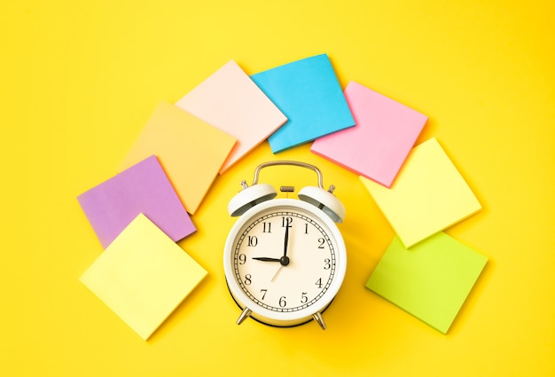 Despertador branco e notas auto-adesivas coloridas em uma tabela amarela. O conceito de tempo de trabalho. Ambiente de trabalho