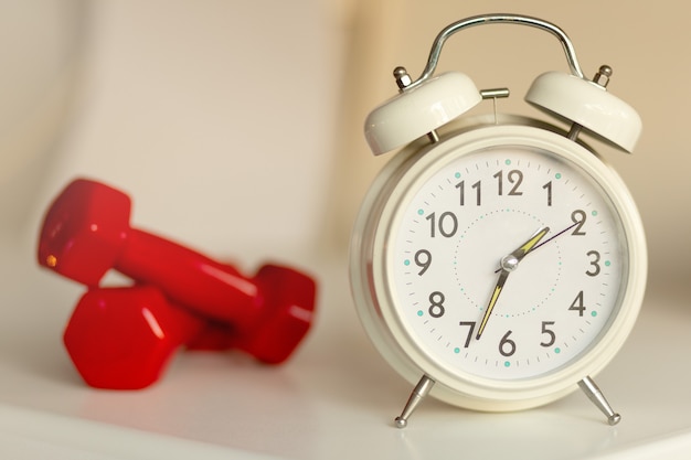 Despertador clásico blanco y pesas rojas desenfocadas sobre fondo blanco Tiempo para hacer ejercicio