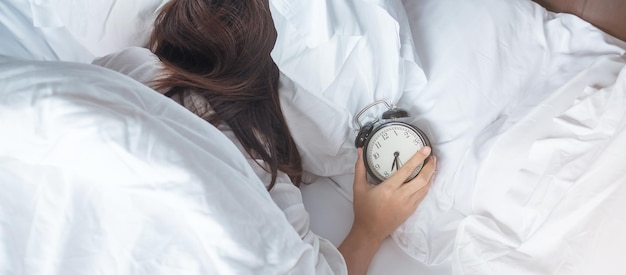 El despertador y la mano de una mujer asiática detienen el tiempo en la cama mientras duermen, una mujer adulta joven se despierta tarde en la mañana. Relajante, somnoliento, rutina diaria y que tengas un buen día conceptos