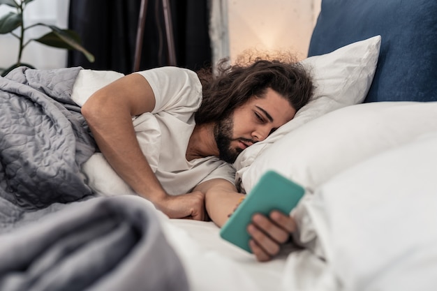 Despertador. moreno com sono olhando para a tela do smartphone enquanto está deitado na cama