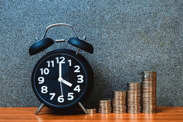 El despertador y el paso de las pilas de las monedas en la tabla de funcionamiento, hora para el concepto del dinero de los ahorros.