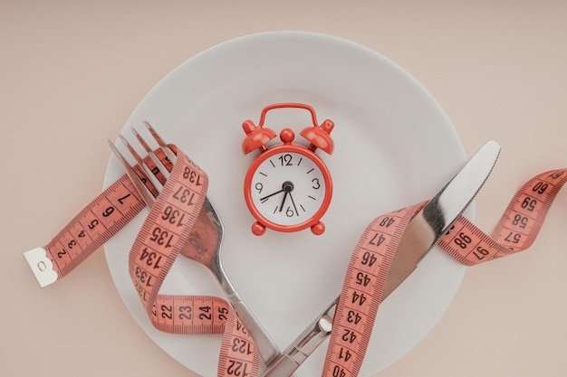 Despertador en plato blanco con cinta métrica y cubiertos. Concepto de dieta de ayuno y pérdida de peso.