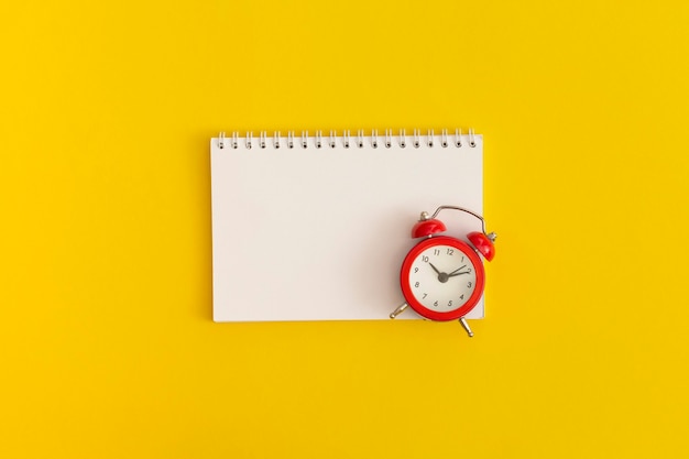 Despertador rojo y cuaderno con espacio para texto sobre fondo amarillo