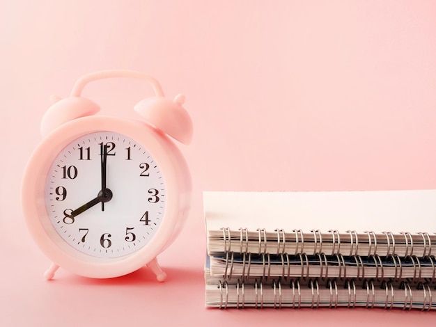 Un despertador rosa a las 8 de la mañana está al lado de una pila de cuadernos sobre un fondo rosa