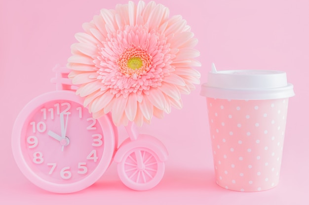 El despertador rosado, el vidrio de café para llevar y el gerbera florecen en fondo rosado.